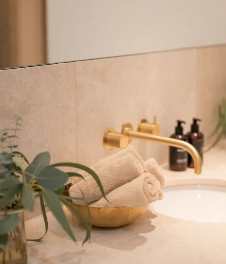 Ein Waschbecken im Almanac Palais Vienna verfügt über einen goldenen Wasserhahn, eine Schale mit aufgerollten beigen Handtüchern, zwei Seifenspender und eine grüne Pflanze auf dem Tresen.